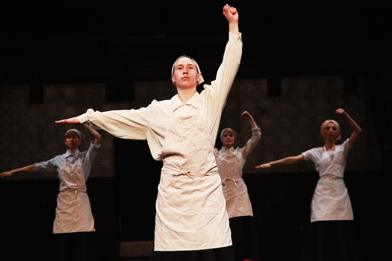 Performers in white aprons and hankerchiefs on their heads with one arm above their heads and another outstretched